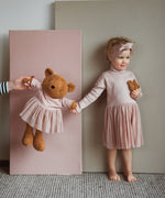 A child standing next to the Wooly Organic soft teddy bear in caramel brown with a pink tutu and holding by the hand. Teddy bear available at Babipur. 