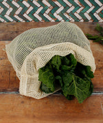 A Slice Of Green large mesh produce bag made from recycled cotton laid flat on a wooden surface showing a bunch of cabbage leaves inside the opened bag showing scale. 