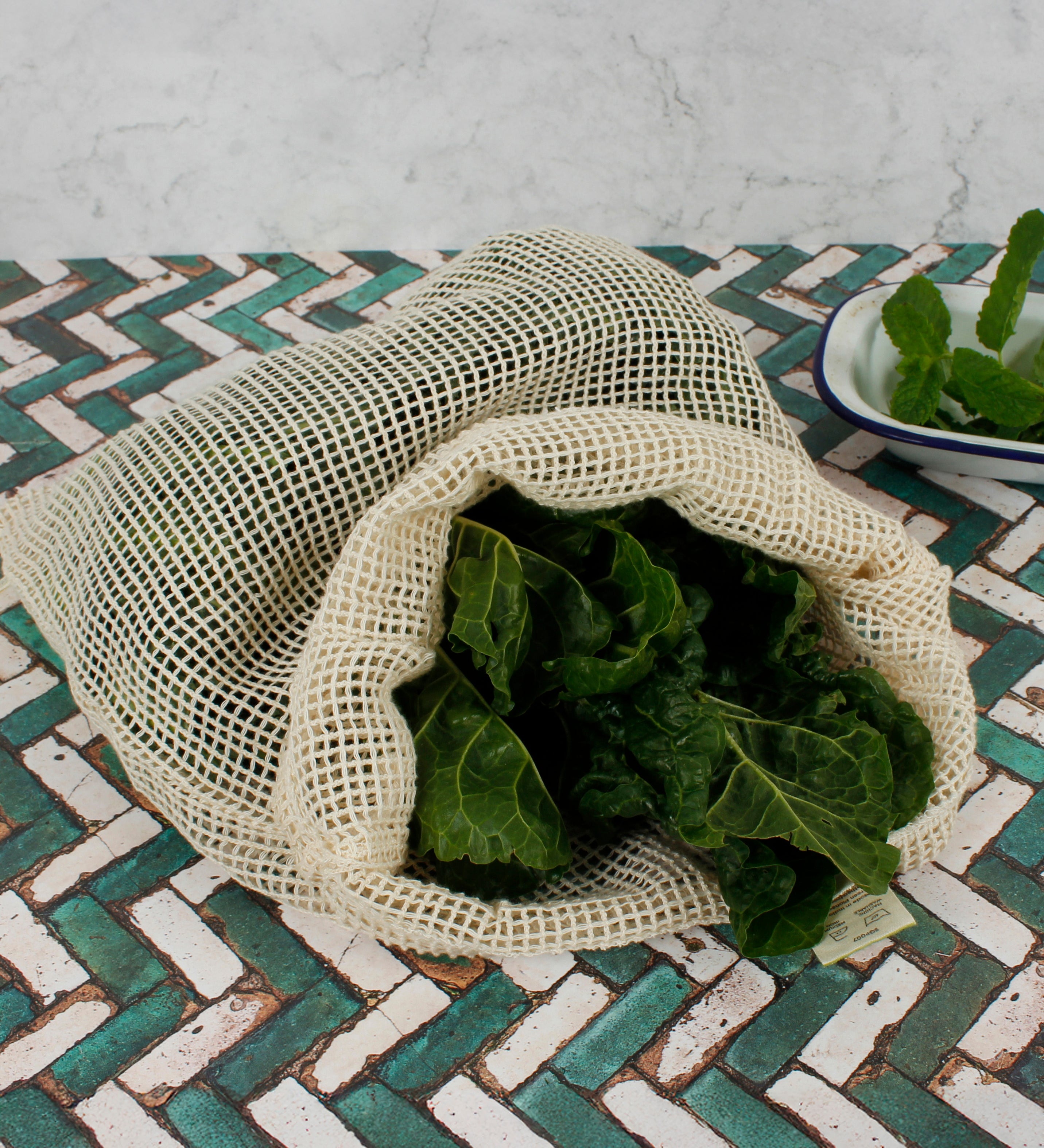A Slice Of Green large mesh produce bag made from recycled cotton on a tiled surface showing a bunch of cabbage leave inside showing scale. 