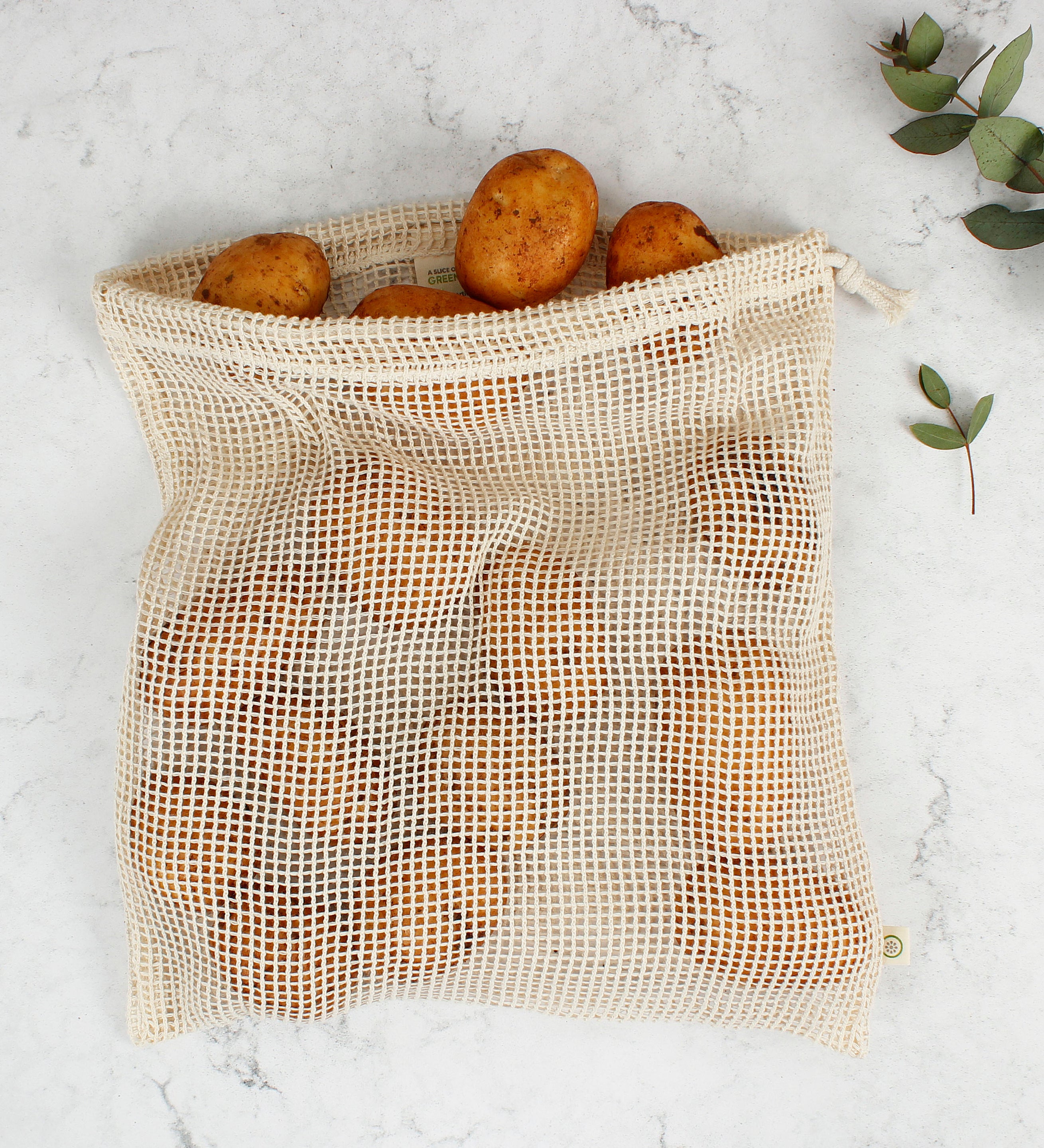 A Slice Of Green large mesh produce bag made from recycled cotton laid flat on a stone surface showing a bunch of potatoes inside showing scale.