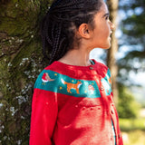 Girl with braided hair wearing a red Frugi organic cotton button-front cardigan with a teal band across the chest decorated with embroidered birds, foxes, and stars.