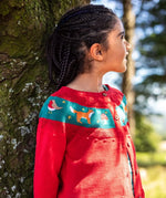 Girl with braided hair wearing a red Frugi organic cotton button-front cardigan with a teal band across the chest decorated with embroidered birds, foxes, and stars.