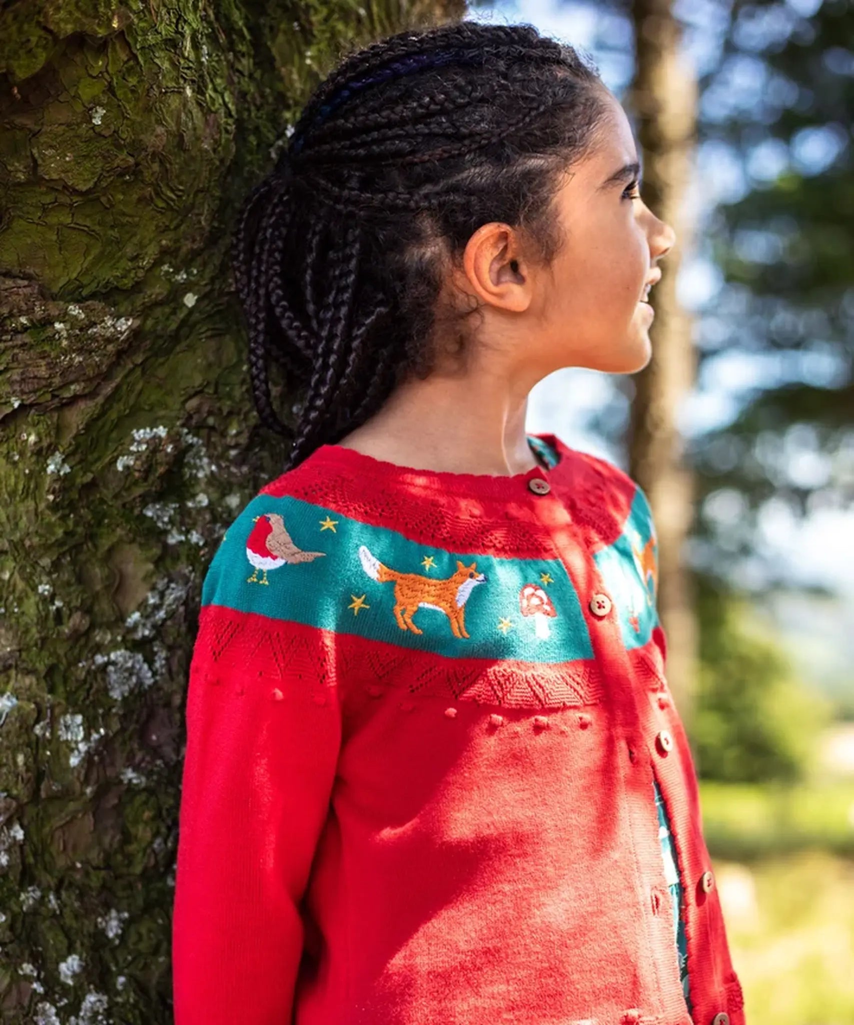Girl with braided hair wearing a red Frugi organic cotton button-front cardigan with a teal band across the chest decorated with embroidered birds, foxes, and stars.