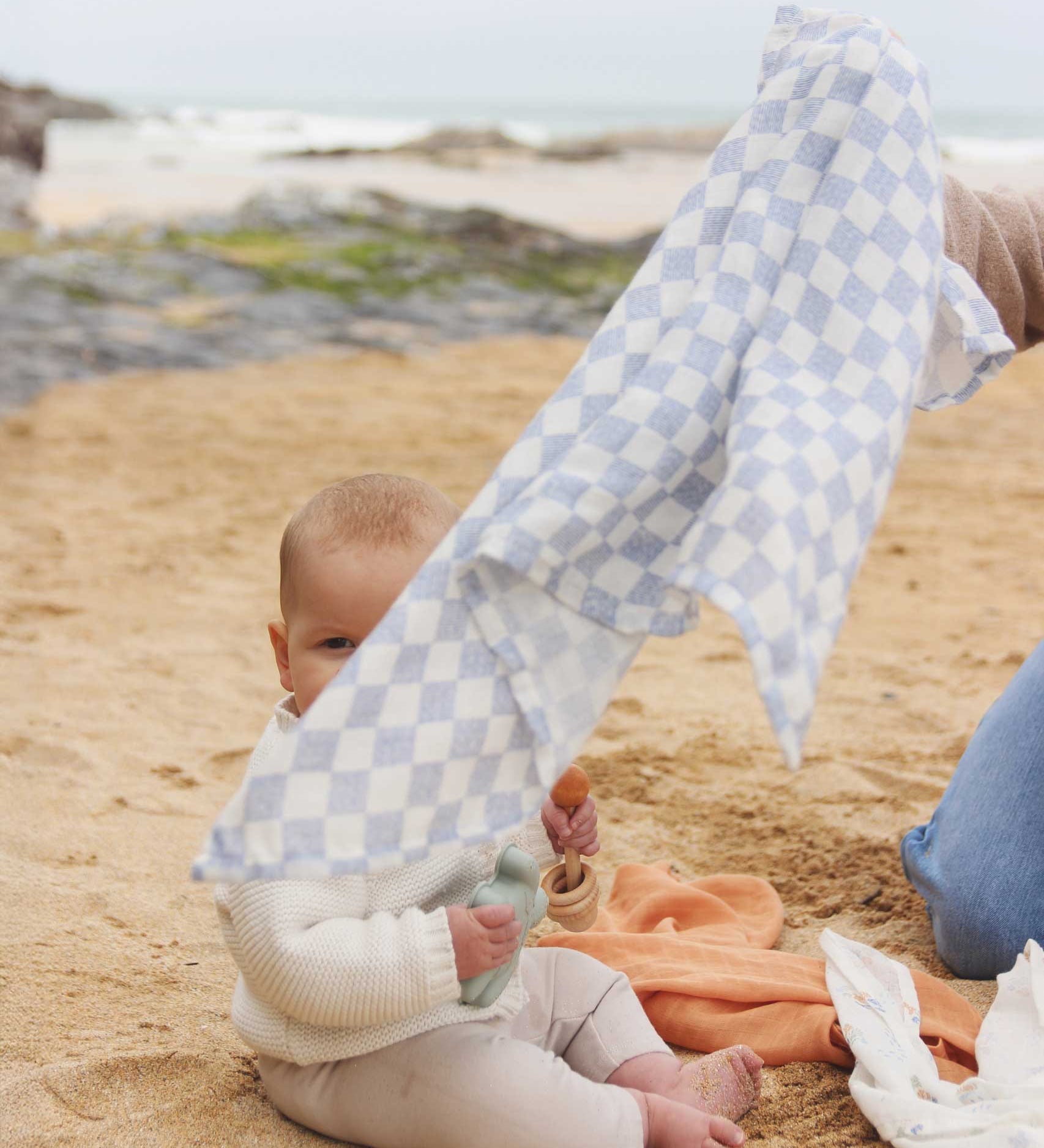 The check blue and white design muslin cloth from the Avery Row Coastline Baby Muslin Squares in a Set being held up by an adults, a baby can be seen sitting behind. The pack has one plain coral coloured cloth, one check blue and white waves design and one coastline print design. Avery Row have a range of stylish baby and nursery essential available here at babipur. 