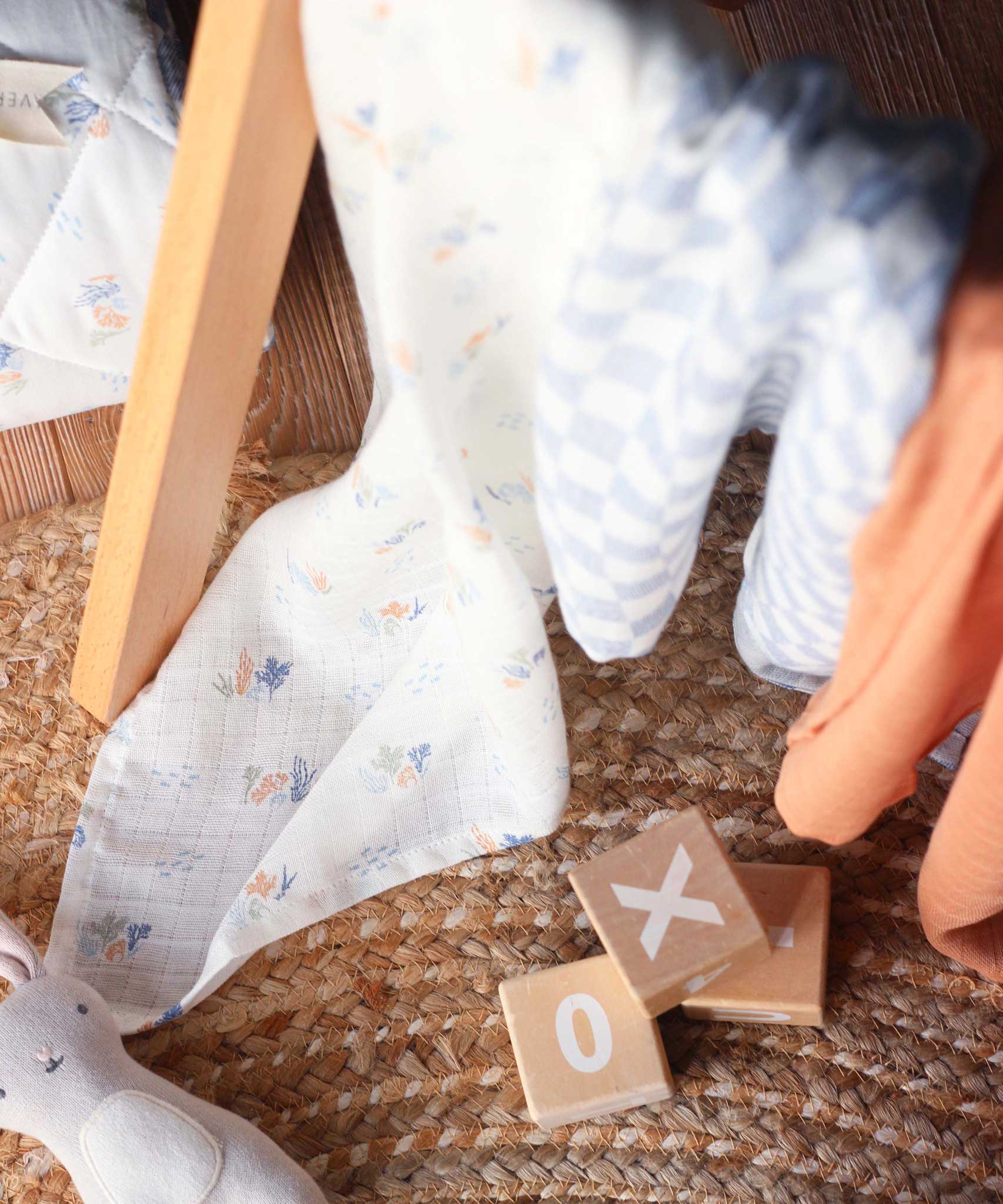 Avery Row Coastline Baby Muslin Squares in a Set of 3. The cloths have been placed on a baby gym. The pack has one plain coral coloured cloth, one check blue and white waves design and one coastline print design. Avery Row have a range of stylish baby and nursery essential available here at babipur. 