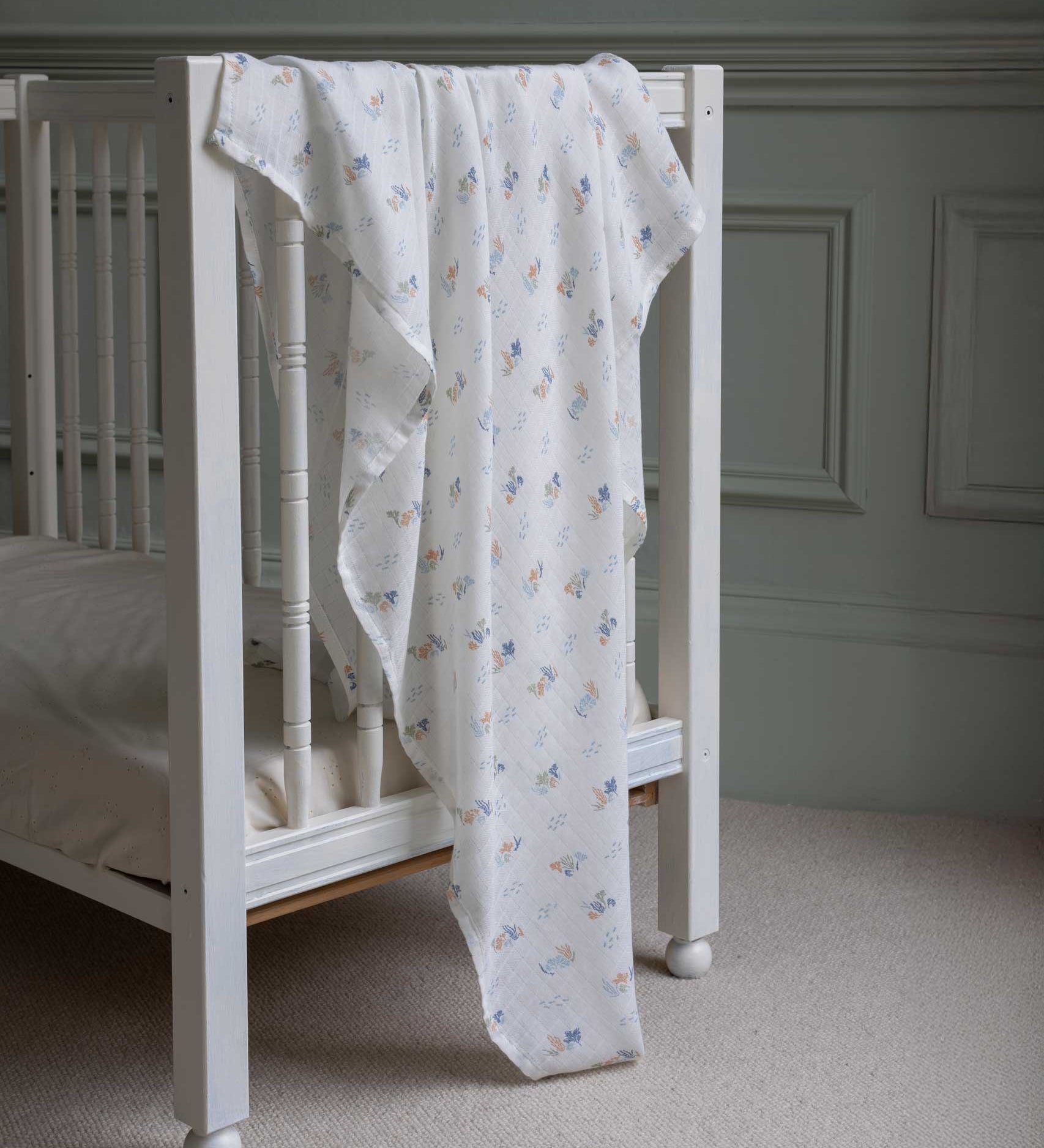 Avery Row Coastline baby Muslin Swaddle. The muslin has been draped over the side of a white cot bed. The swaddle features a delicate coastline print that has corals and little blue fish. Avery Row have a collection of stylish nursery and baby essentials available at Babipur.