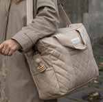 A close up of a person carrying the Avery Row Baby Changing Bag in a  Natural colour on their shoulder with the shoulder strap. A wooden toy can be seeing in the side pocket