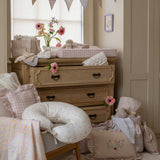 An Avery Row Nursing Pillow with an Anemone design placed on a chair in a nursery. The The top of the pillow features a white background with a delicate pink anemone flower print. Avery Row have a range of baby and nursery products with stylish designs available at Babipur. 