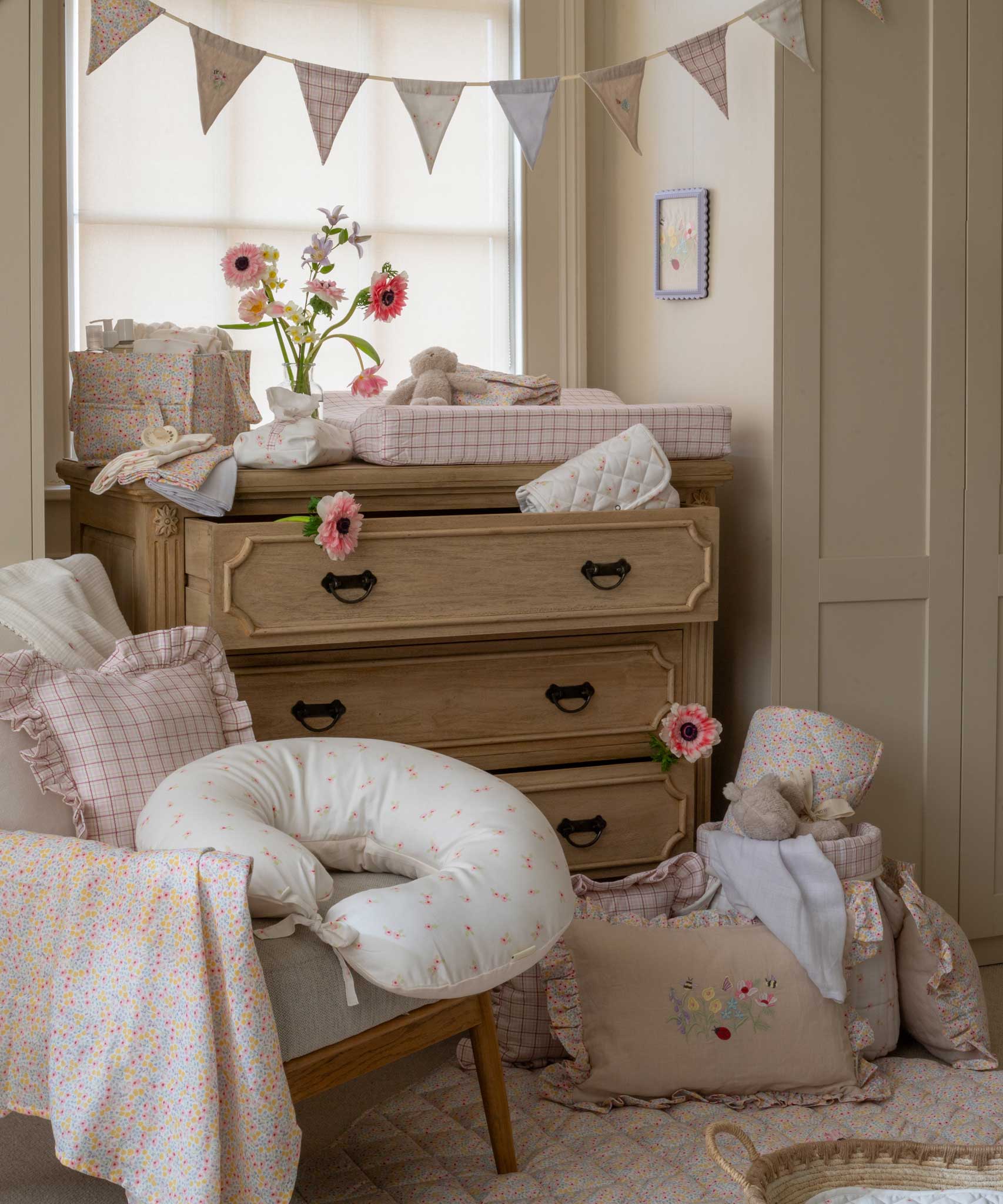 An Avery Row Nursing Pillow with an Anemone design placed on a chair in a nursery. The The top of the pillow features a white background with a delicate pink anemone flower print. Avery Row have a range of baby and nursery products with stylish designs available at Babipur. 