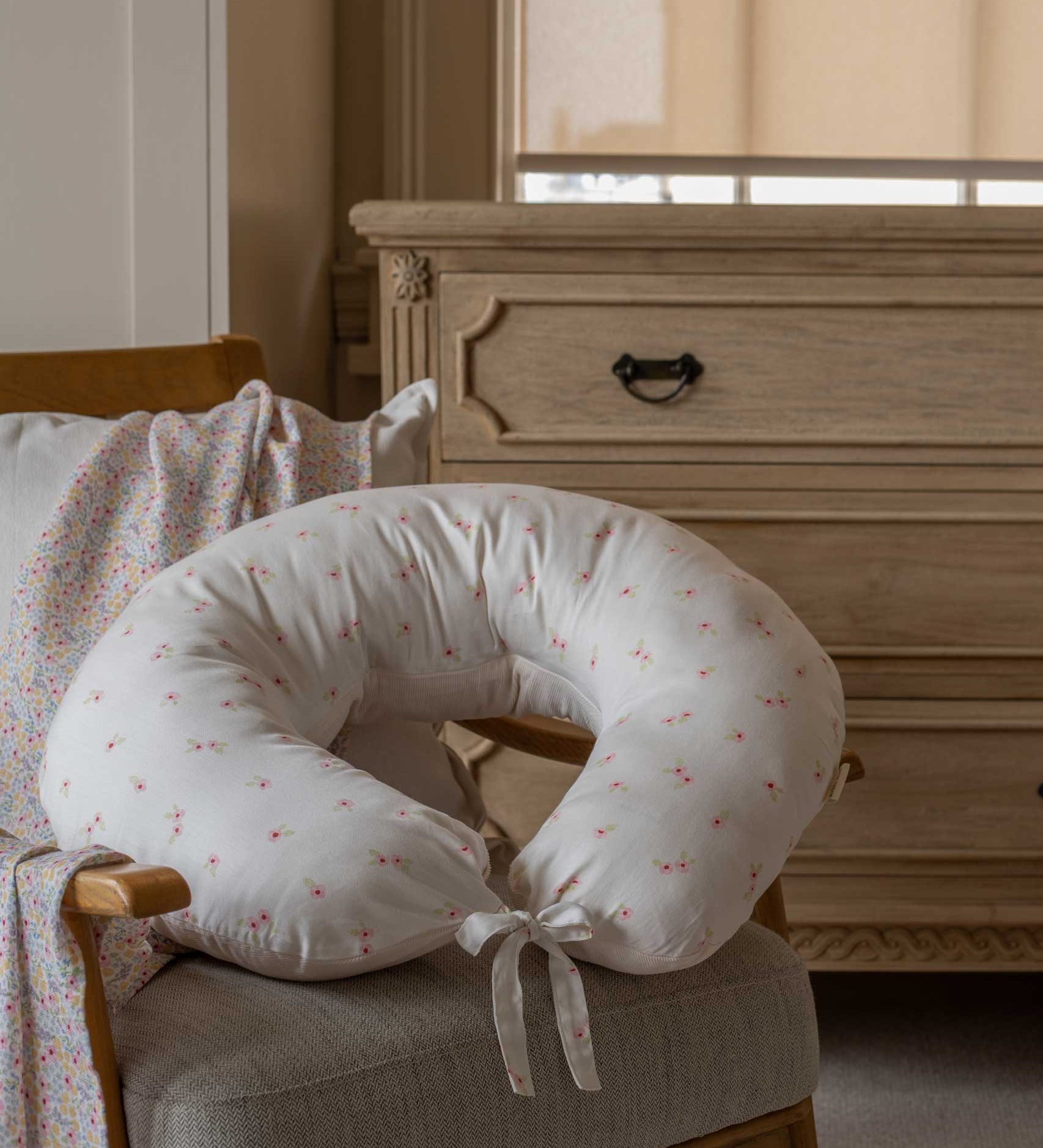 An Avery Row Nursing Pillow with an Anemone design placed on a chair in a nursery. The The top of the pillow features a white background with a delicate pink anemone flower print. Avery Row have a range of baby and nursery products with stylish designs available at Babipur. 