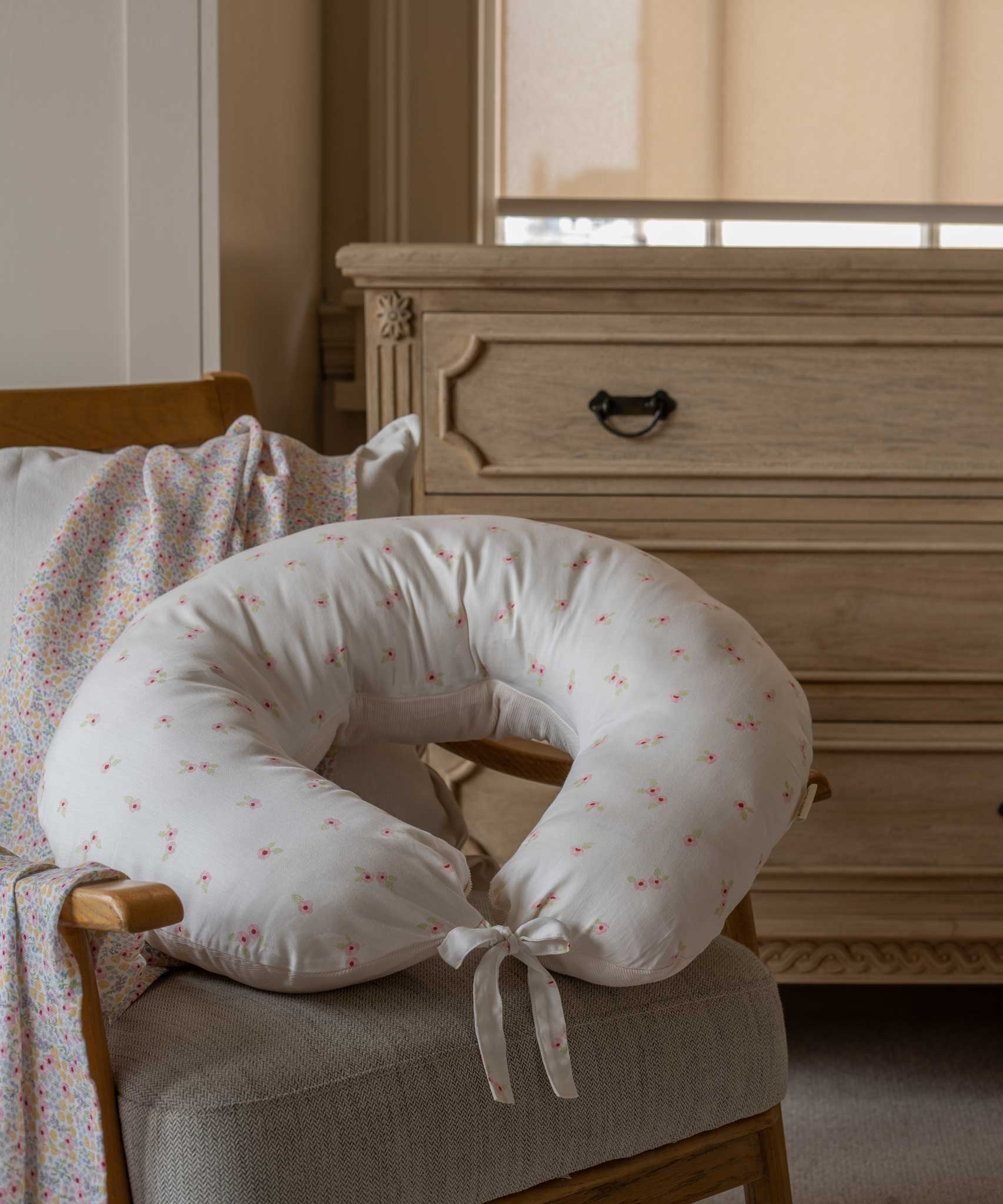 An Avery Row Nursing Pillow with an Anemone design placed on a chair in a nursery. The The top of the pillow features a white background with a delicate pink anemone flower print. Avery Row have a range of baby and nursery products with stylish designs available at Babipur. 