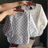 An adults hand holding a hanger that has an Avery Row Sleeved Bib with a check Waves design hung on it. Another design can be seen in the background. This blue and white coloured bib has long sleeves with elasticated cuffs and features a pocket to help catch any food debris. We have a wide range of Avery Row stylish baby products available at Babipur. 