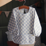 An adults hand holding a hanger that has an Avery Row Sleeved Bib with a check Waves design hung on it. This blue and white coloured bib has long sleeves with elasticated cuffs and features a pocket to help catch any food debris. We have a wide range of Avery Row stylish baby products available at Babipur. 