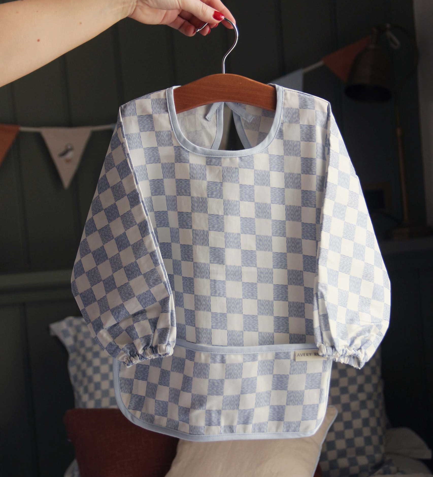 An adults hand holding a hanger that has an Avery Row Sleeved Bib with a check Waves design hung on it. This blue and white coloured bib has long sleeves with elasticated cuffs and features a pocket to help catch any food debris. We have a wide range of Avery Row stylish baby products available at Babipur. 