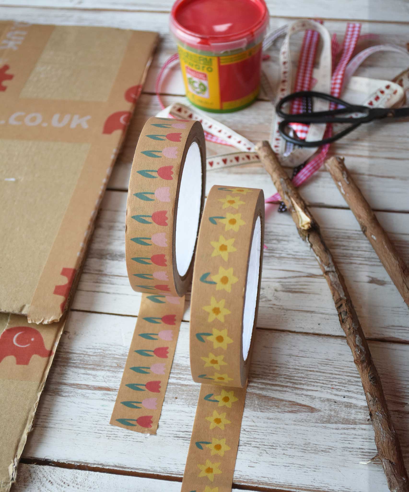 A roll of tulip and a roll of daffodils kraft paper tape from Babipur with a bit rolled out on each and placed on a wooden surface. Craft supplies can be seen in the background. 