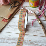 A roll of pink tulip printed kraft paper tape from Babipur with a bit rolled out and placed on a wooden surface. Craft supplies can be seen in the background including some red Okonorm finger paint.. 