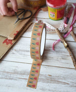 A roll of pink tulip printed kraft paper tape from Babipur with a bit rolled out and placed on a wooden surface. Craft supplies can be seen in the background including some red Okonorm finger paint.. 