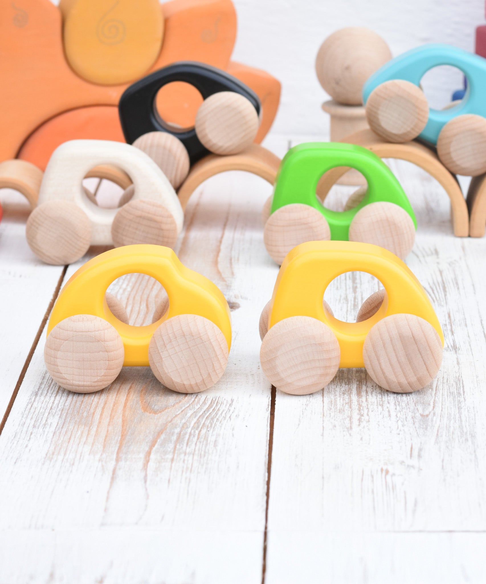 A Bajo handmade wooden yellow mini car next to a beetle car available at Babipur. Photo is in a playroom setting with Grimms blocks in the background and other Bajo mini cars in the background.