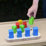 Bajo 10 piece sorting board. Wooden triangle base with 10 slots, 4 square, 3 triangle, 2 rectangle and 1 circle with 10 blocks to fit inside. Child's hand placing a green triangle block onto the base.
