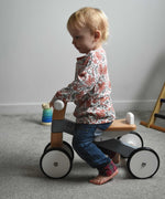 A child sitting on a Bajo Toys wooden Cycle toddler Ride-On in the monochrome, white and grey colourway. The wooden ride on cycle is part of a wode range of wooden toys available here at Babipur. 