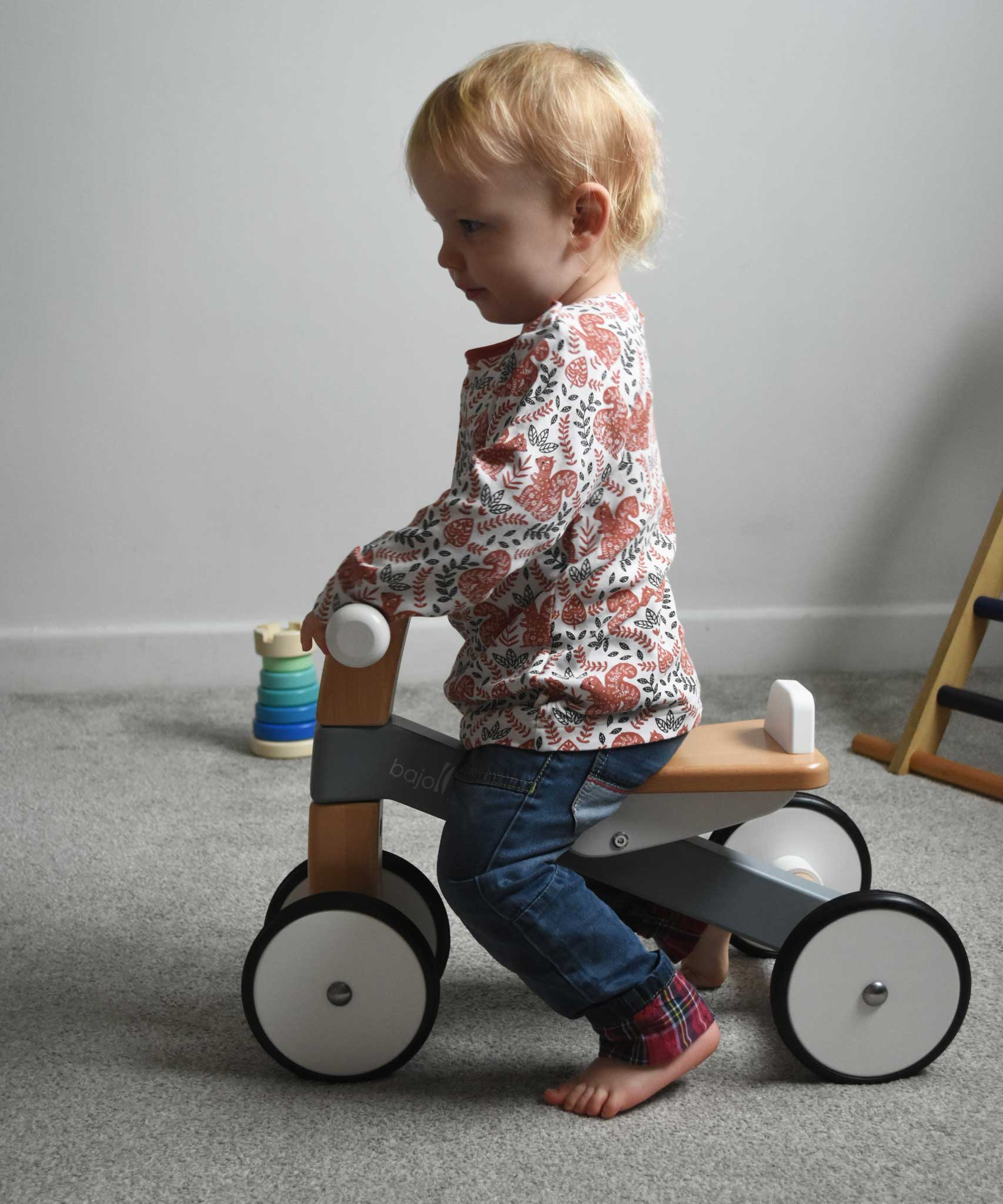 A child sitting on a Bajo Toys wooden Cycle toddler Ride-On in the monochrome, white and grey colourway. The wooden ride on cycle is part of a wode range of wooden toys available here at Babipur. 