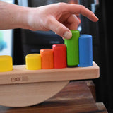 A close up of an adult's hand placing blocks on the base of the Bajo Balance Scale wooden toy. A wooden base with a curved base with coloured wooden blocks that sit on top. These heirloom quality toys by Bajo are part of a wide range of wooden toys available here at Babipur.