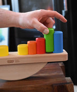 A close up of an adult's hand placing blocks on the base of the Bajo Balance Scale wooden toy. A wooden base with a curved base with coloured wooden blocks that sit on top. These heirloom quality toys by Bajo are part of a wide range of wooden toys available here at Babipur.