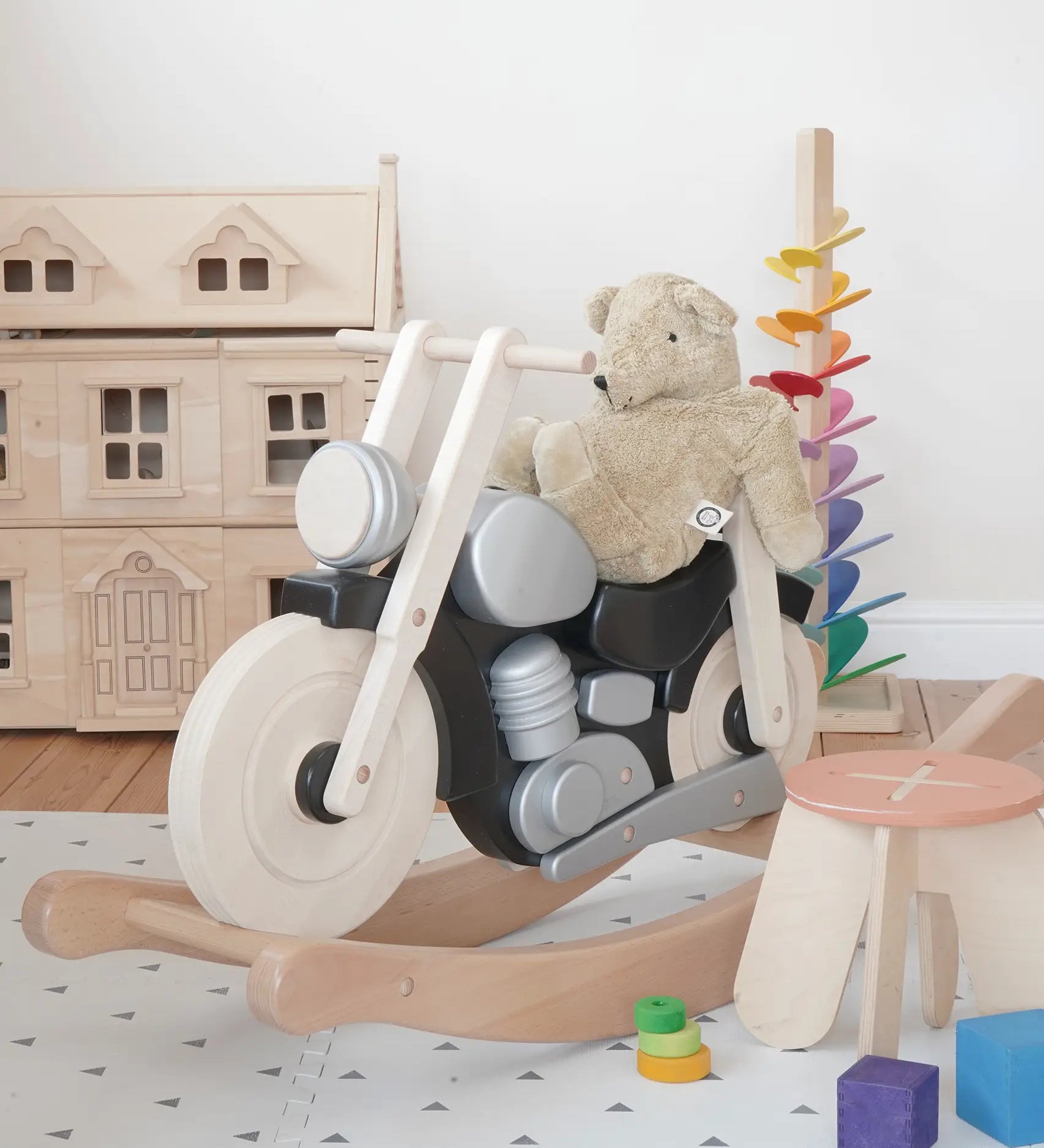 Bajo wooden black rocking bike with a senger teddy bear on top showing scale with a doll house and marble tree in the background
