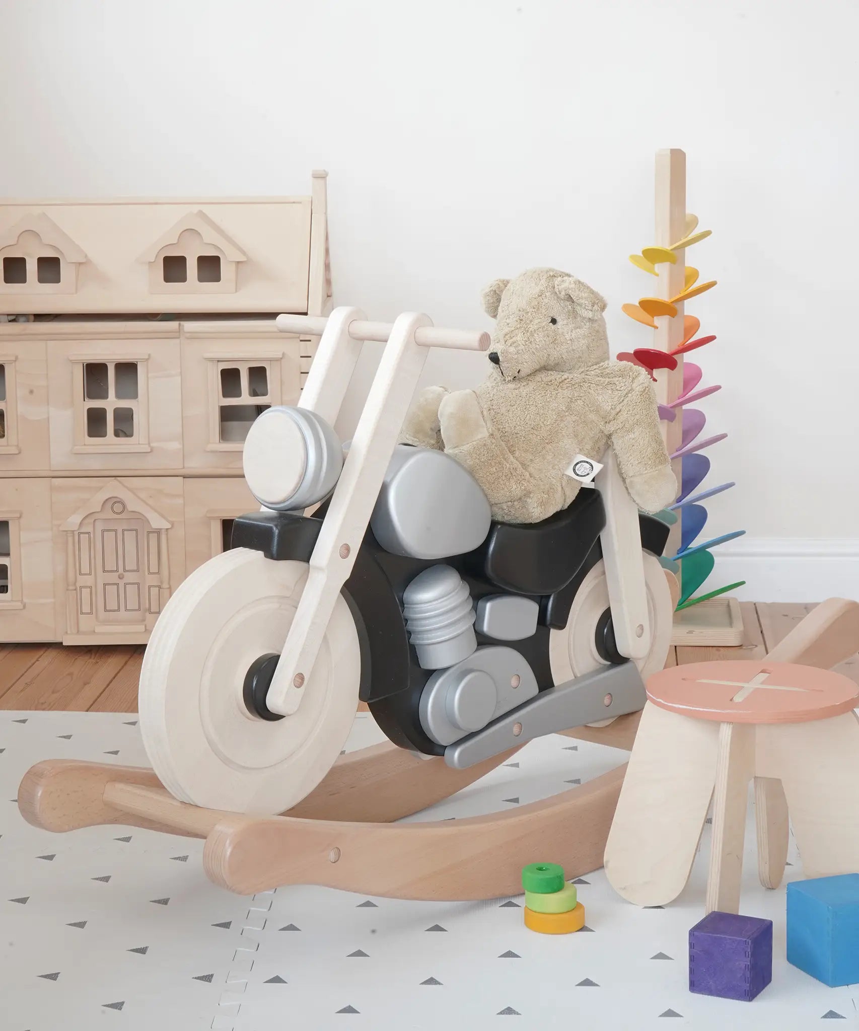 Bajo wooden black rocking bike with a senger teddy bear on top showing scale with a doll house and marble tree in the background