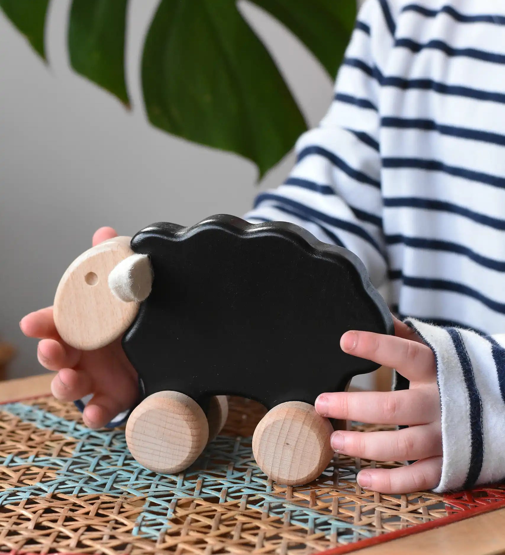 Child reaching for the Bajo black sheep push along wooden toy on wheels placed a rattan stool in the Babipur playroom.