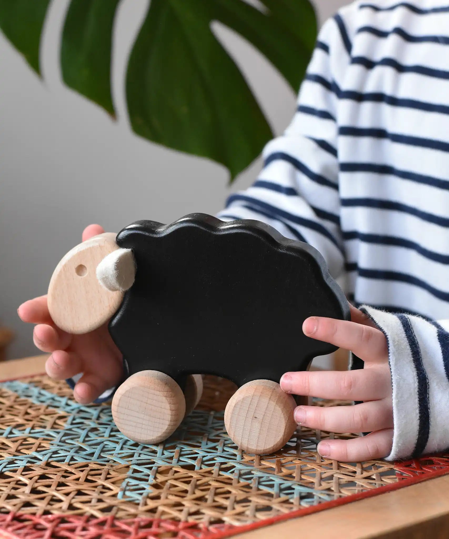 Child reaching for the Bajo black sheep push along wooden toy on wheels placed a rattan stool in the Babipur playroom.