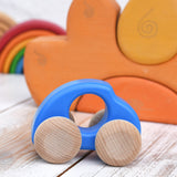 A blue Bajo handmade wooden beetle style car available at Babipur. Vehicle has unpainted wheels and can be pulled or pushed along. Photo is in a playroom setting with a Grimms rainbow and sun block sets in the background