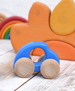 A blue Bajo handmade wooden beetle style car available at Babipur. Vehicle has unpainted wheels and can be pulled or pushed along. Photo is in a playroom setting with a Grimms rainbow and sun block sets in the background