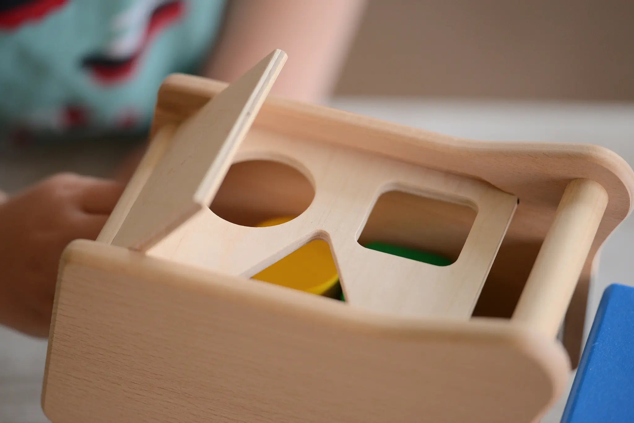 Close-up of a wooden shape-sorting toy box on the Bajo blue tipper truck shape sorter with a hinged lid, showing circular, rectangular, and triangular cutouts, with colorful blocks partially visible inside.