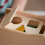 Close-up of a wooden shape-sorting toy box on the Bajo blue tipper truck shape sorter with a hinged lid, showing circular, rectangular, and triangular cutouts, with colorful blocks partially visible inside.