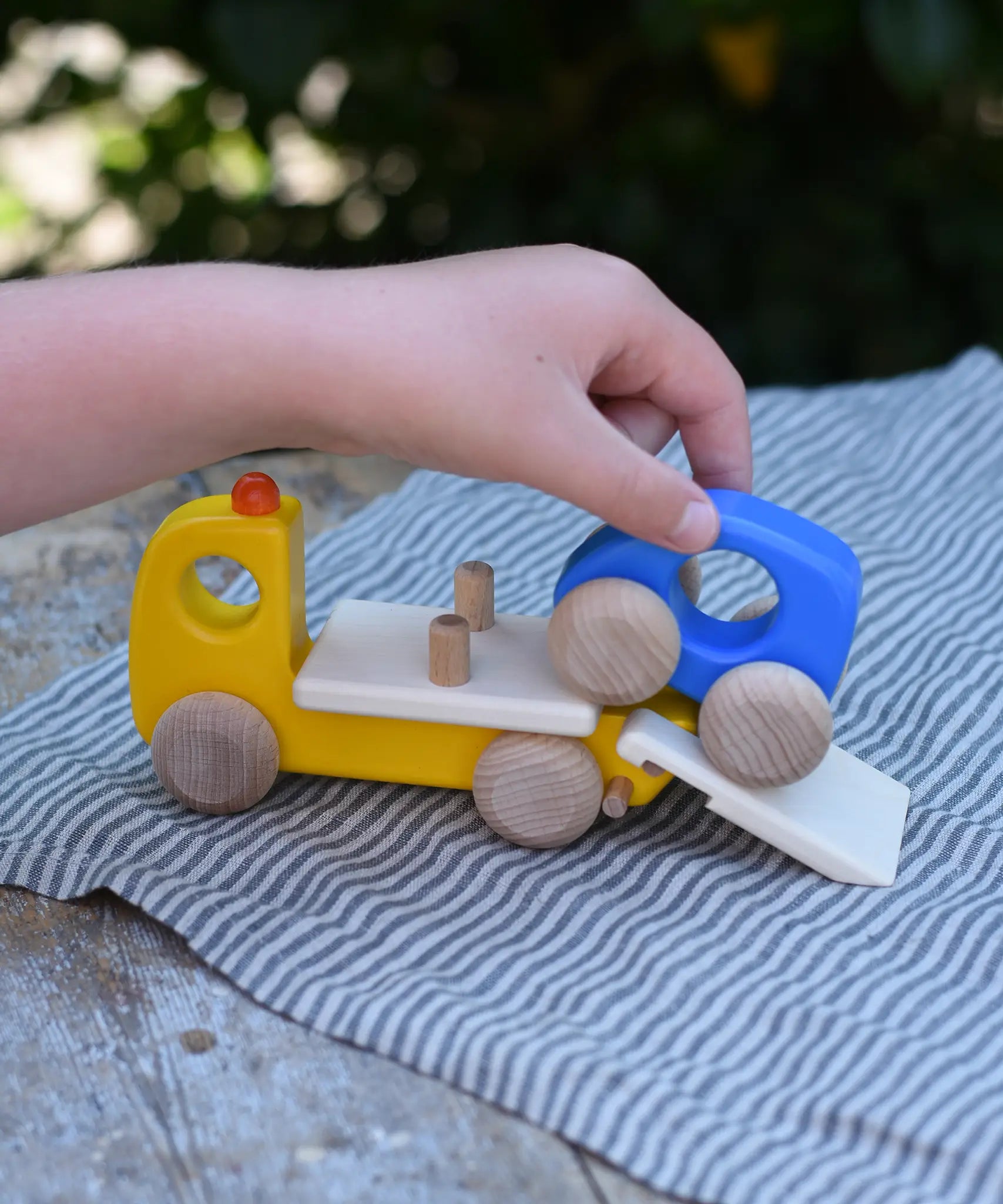 Bajo yellow breakdown lorry wooden toy with a child's hand driving the blue car up the ramp onto the trailer.