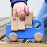 A close up of a child playing with a blue coloured Bajo trucks with blocks, the child's hand in covered in soil and lifting one of the blocks from the back of the truck