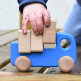 A close up of a child playing with a blue coloured Bajo trucks with blocks, the child's hand in covered in soil and lifting one of the blocks from the back of the truck
