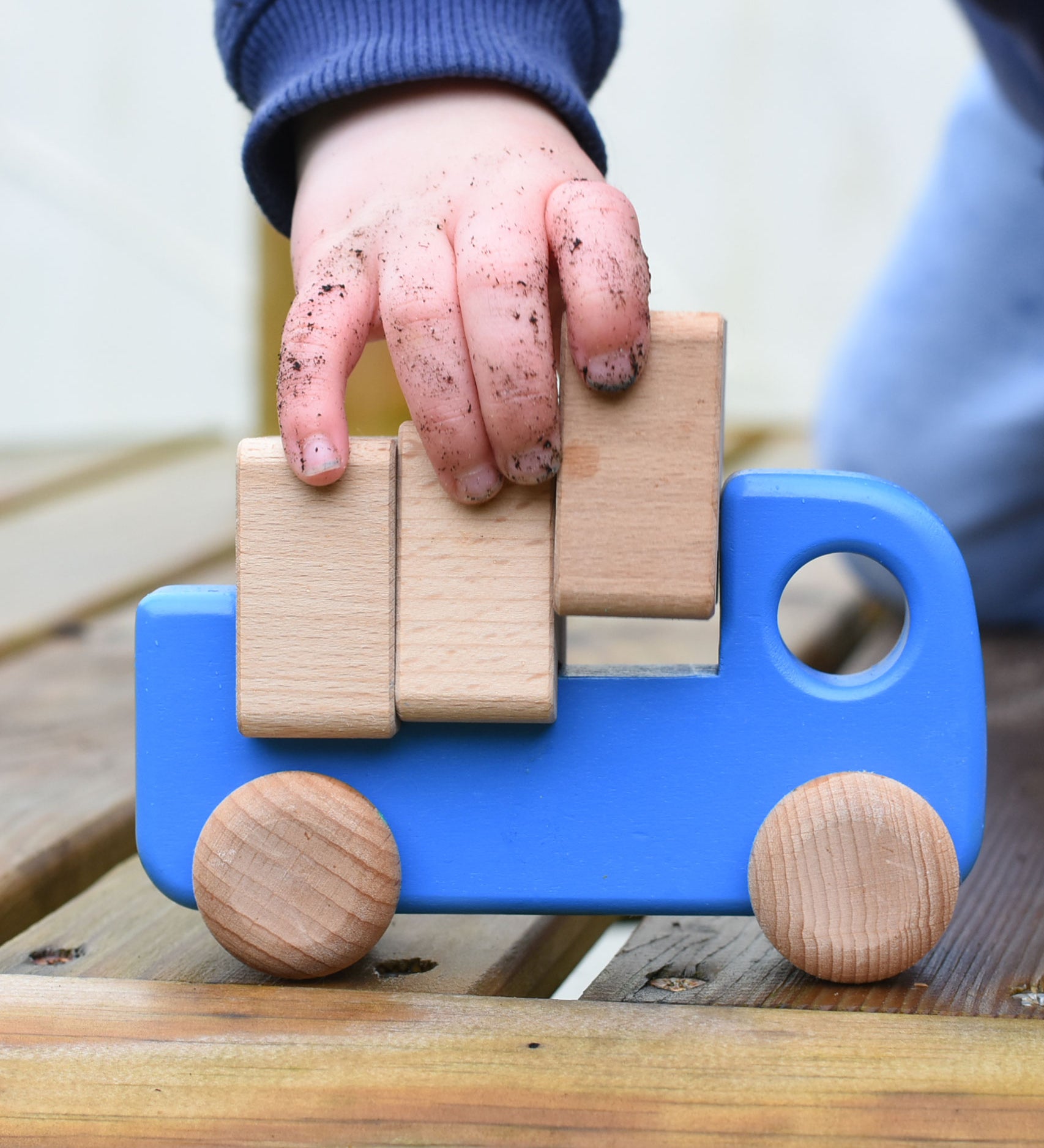 A close up of a child playing with a blue coloured Bajo trucks with blocks, the child's hand in covered in soil and lifting one of the blocks from the back of the truck.  This wooden truck is part of a wide range of Bajo wooden toy vehicles available at Babipur.