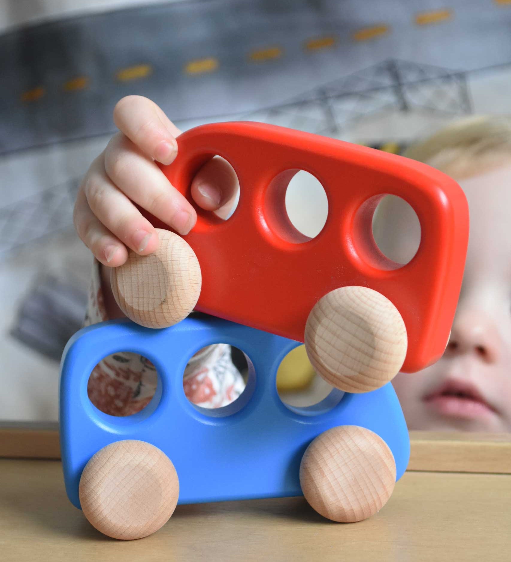 A child's hand holding a red coloured bus, the child is placing it on a blue coloured bus. These heirloom quality toys by Bajo are part of a wide range of wooden toys available here at Babipur. 