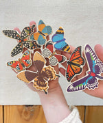 An adult holding all the butterfly shaped pieces from the Bajo World Of Butterflies Sorter Puzzle in their hand. This puzzle has butterfly shaped pieces that are decorated on both sides that slot into the coordinating shapes on the base. These heirloom quality toys by Bajo are part of a wide range of wooden toys available here at Babipur.