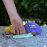 Bajo yellow car and purple camper wooden toy vehicle with green surfboard piece placed in front of it, a child's hand is reaching for the surf board. 