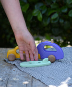 Bajo yellow car and purple camper wooden toy vehicle with green surfboard piece placed in front of it, a child's hand is reaching for the surf board. 