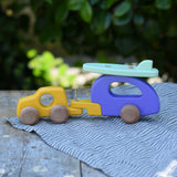 Bajo yellow car and purple camper wooden toy vehicle with green surfboard placed on a striped cloth on a wooden table outdoors.