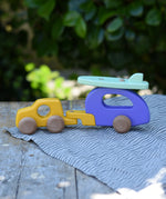 Bajo yellow car and purple camper wooden toy vehicle with green surfboard placed on a striped cloth on a wooden table outdoors.