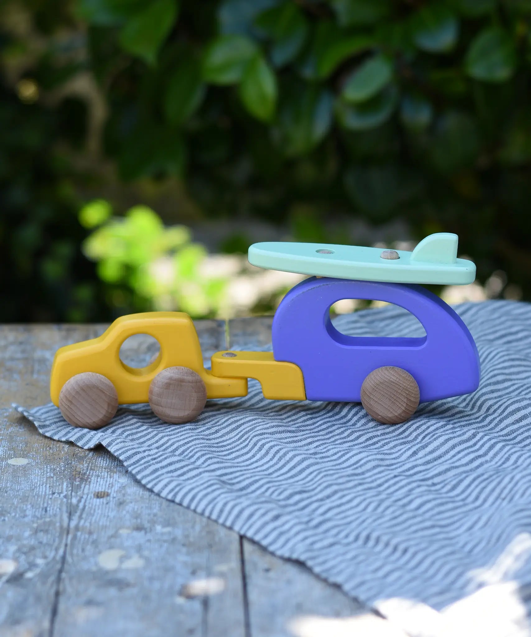 Bajo yellow car and purple camper wooden toy vehicle with green surfboard placed on a striped cloth on a wooden table outdoors.