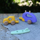 Bajo yellow car and purple camper wooden toy vehicle with green surfboard with all pieces shown separated and placed on a wooden table outdoors.