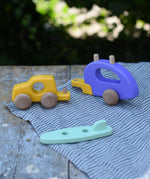 Bajo yellow car and purple camper wooden toy vehicle with green surfboard with all pieces shown separated and placed on a wooden table outdoors.