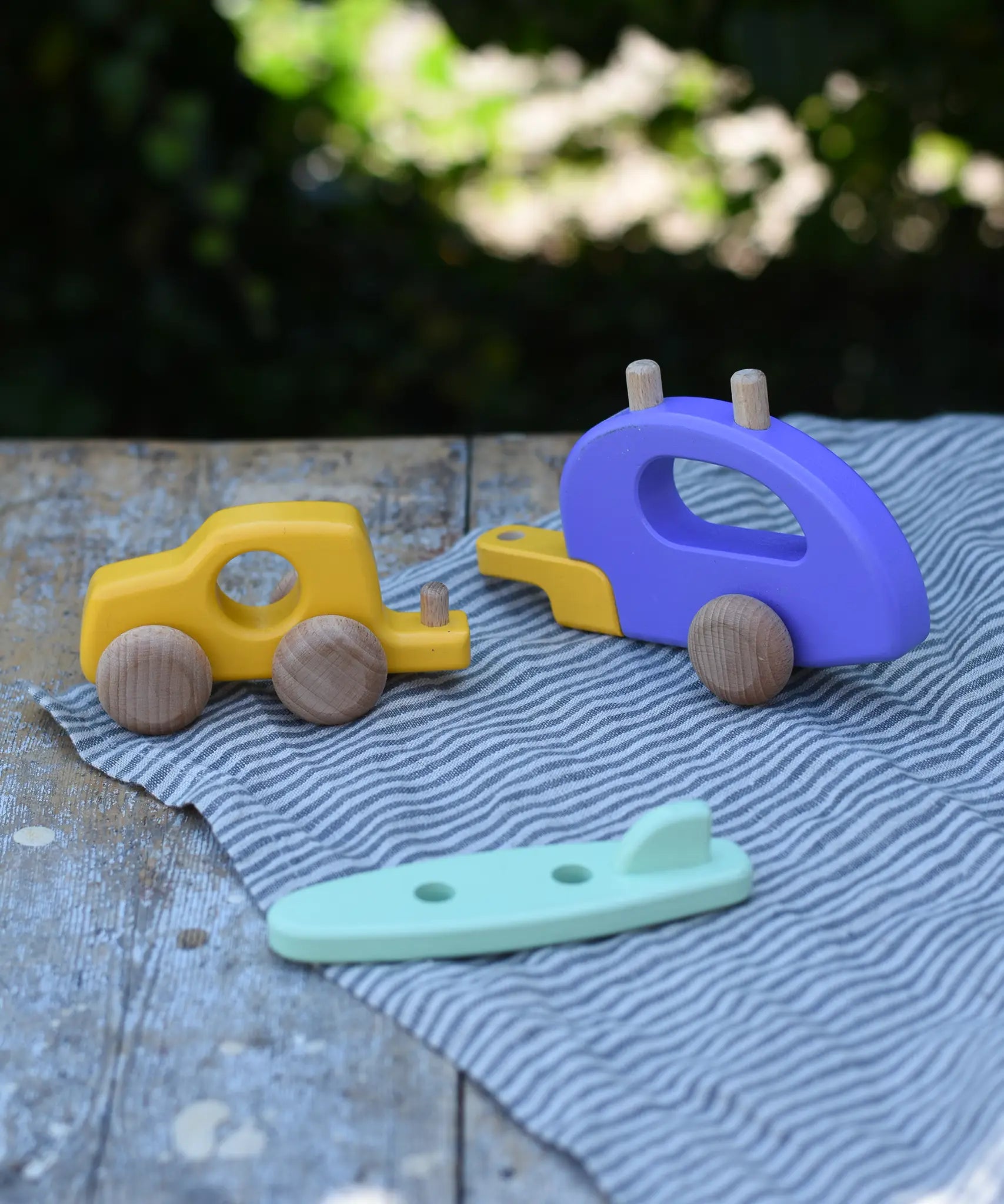 Bajo yellow car and purple camper wooden toy vehicle with green surfboard with all pieces shown separated and placed on a wooden table outdoors.