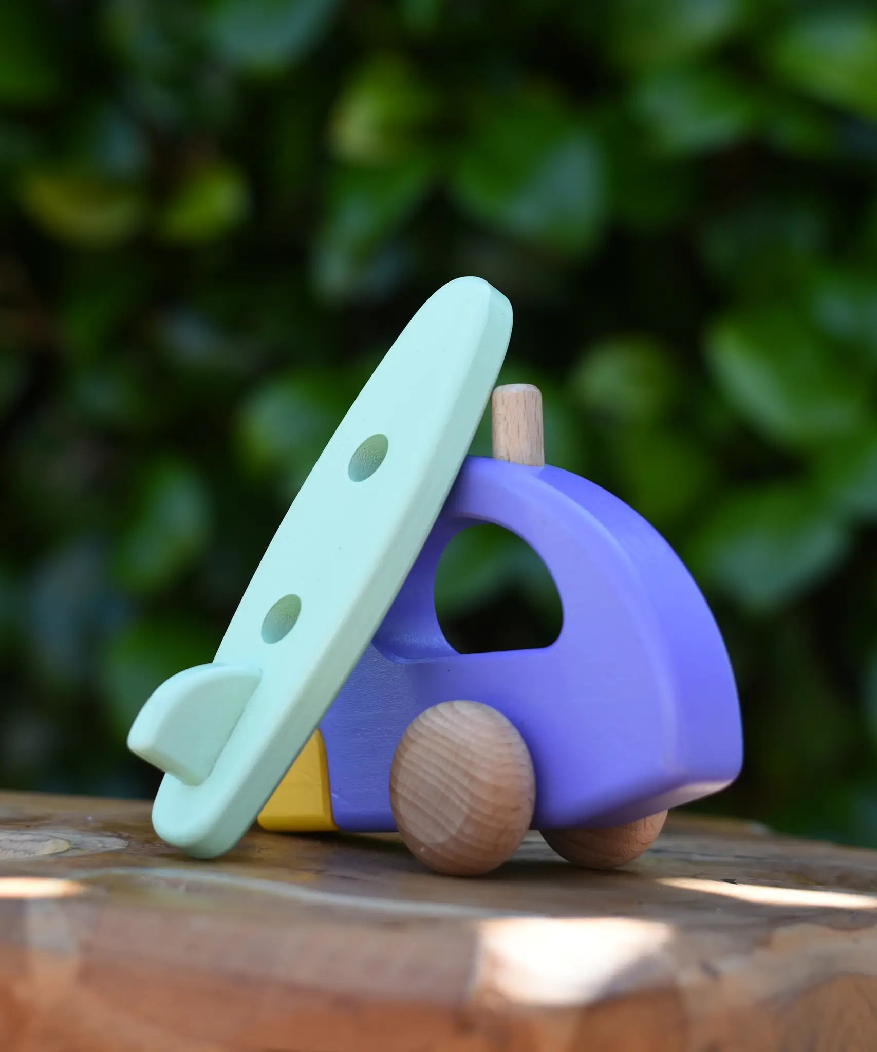 Green surfboard from the Bajo yellow car and purple camper wooden toy vehicle set resting on the camper.